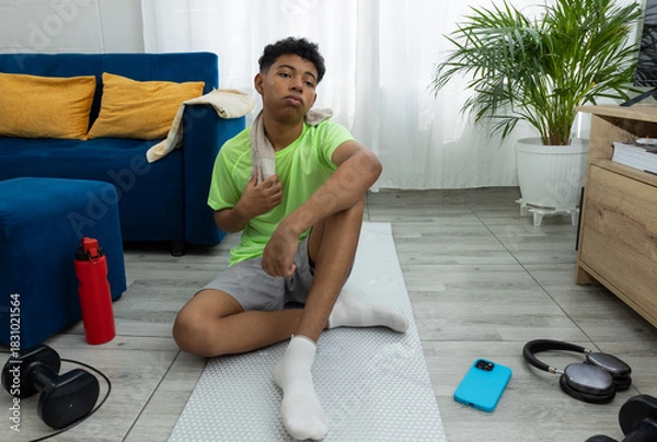Obraz Brown skinned teenager sitting on an exercise mat, sweating and looking tired while resting with a towel around his neck. A scene of home workout and recovery