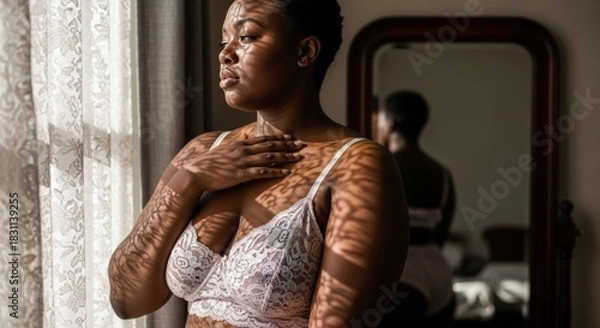 Fototapeta An African American woman in a white lace bra stands by a window. Sunlight casts intricate shadow patterns on her skin, with a blurred mirror reflection behind her.