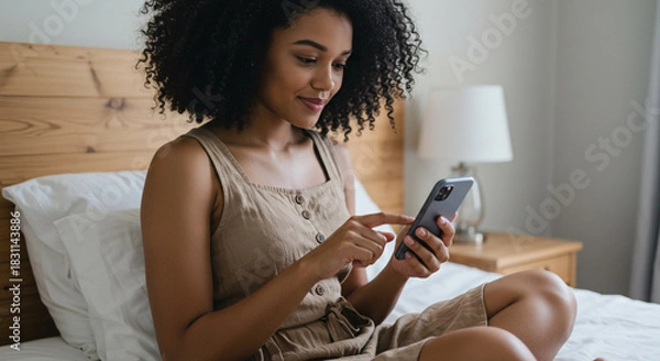 Fototapeta Smiling woman browsing smartphone, relaxing comfortably on a cozy bed, enjoying modern connectivity and leisure time