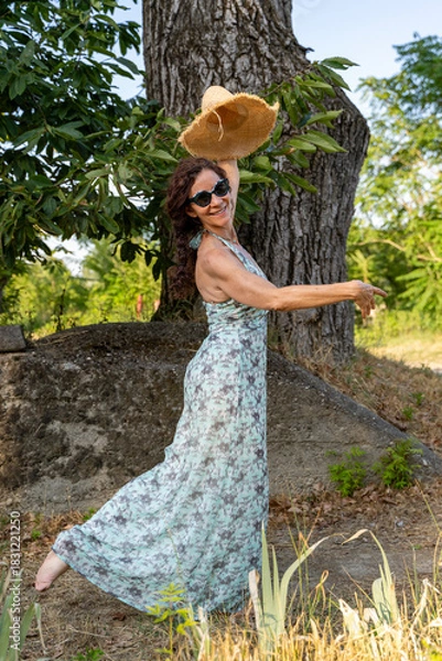 Fototapeta Woman playing with straw hat and dancing barefoot with a big smile in nature