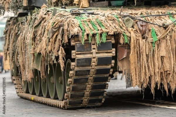 Fototapeta Close-up view of a military tracked tank covered in camouflage netting, showing its heavy armored treads and weathered fabric strips