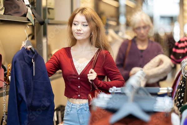 Obraz In sales area of store, young woman customer chooses color and style of sweater. Creating capsule wardrobe. Outlet, discount day on Black Friday. Buyer enjoys shopping