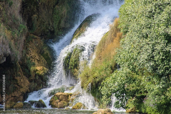 Obraz A close-up view of the Kravica Waterfall in Herzegovina, showing cascading water over moss-covered rocks surrounded by dense green vegetation.