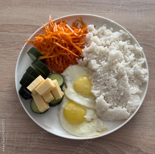 Obraz A simple meal features rice, two fried eggs, fresh cucumbers, and grated carrot salad on a plate