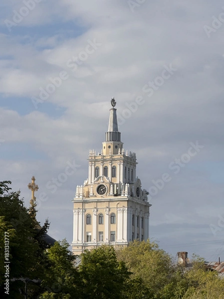 Obraz A tall clock tower with intricate design stands prominently above surrounding trees on a cloudy day