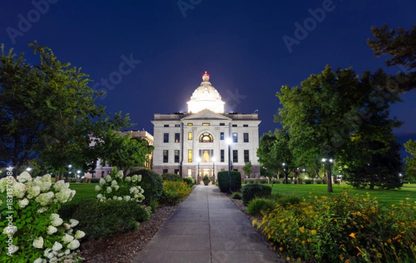 Fototapeta Illuminated South Dakota State Capitol building in Pierre at dusk. Walkway with flowers leads to the building, framed by trees under a clear blue sky