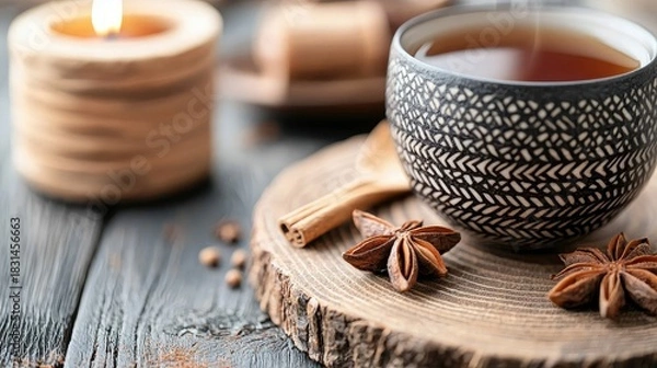 Fototapeta A cup of tea with spices is placed on a wooden board, with a lit candle in the background, creating a cozy and relaxing atmosphere.