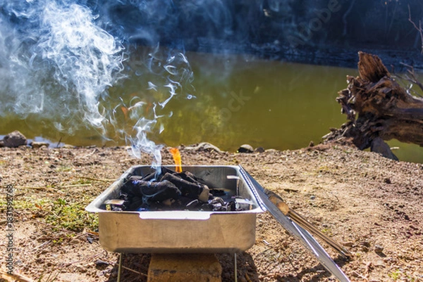 Obraz Portable grill with burning charcoal and smoke by riverbank.