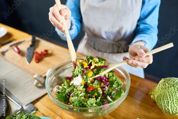 Fototapeta Close-up shot of unrecognizable housewife wearing denim shirt and apron standing at kitchen table and mixing appetizing salad