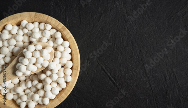 Obraz Organic tapioca pearls in wooden bamboo bowl on black table top view with copy space. Spoon scooping white dry tapioca pearls, cooking ingredient, gluten free food, healthy eating.