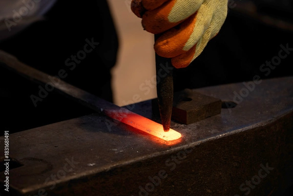 Fototapeta Doha, Qatar - December 01, 2025: Close up of a blacksmith shaping a glowing hot metal piece on an anvil, using a chisel while wearing protective gloves.