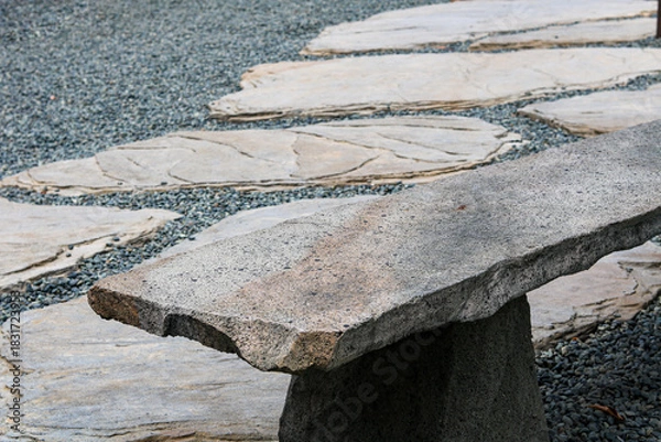 Fototapeta A view of a natural stone bench, crafted from rough-cut slab, overlooking a tranquil garden path of flagstones and dark grey gravel.