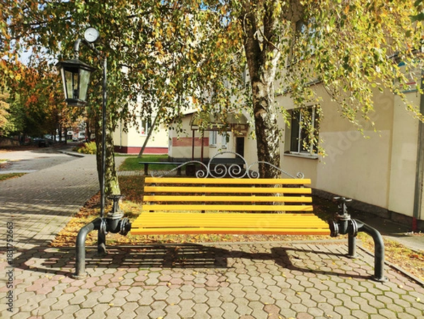 Obraz Yellow park bench with decorative metalwork next to a street lamp featuring a clock, under a birch tree with autumn leaves on a sunny day in an urban setting