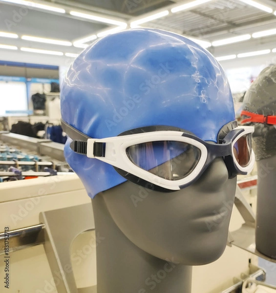Fototapeta Mannequin head display of blue swimming cap and white swimming goggles in a retail store setting, showcasing essential swimwear accessories for sport and recreation