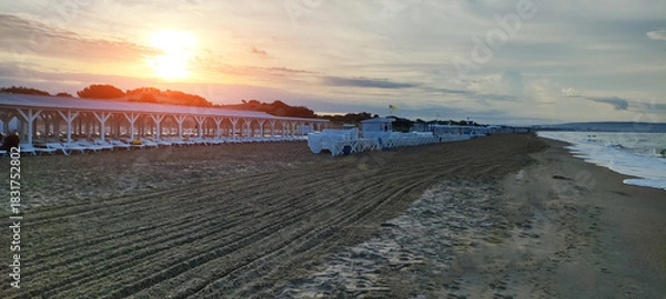 Fototapeta Beach scene at sunset with rows of white sun loungers under white wooden pergola, groomed sand patterns, ocean waves approaching shore, and warm sky colors.