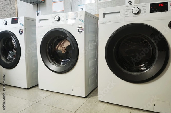 Fototapeta Row of three modern white washing machines with black doors and control panels in laundry room interior. Appliances on tiled floor, ready for use or display.