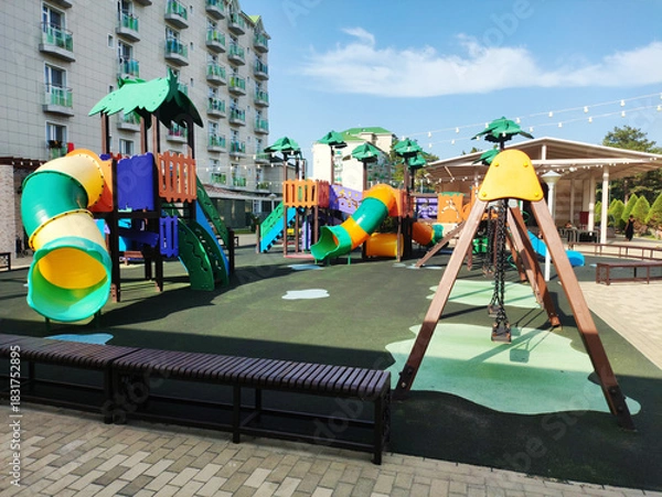 Fototapeta Colorful playground equipment with multiple slides and swing sets on soft rubberized ground surface next to modern multistory building under clear blue sky