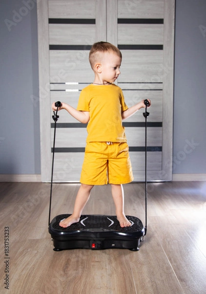 Obraz Handsome 3 year old boy in yellow suit sits on modern vibration platform for exercising at home, background