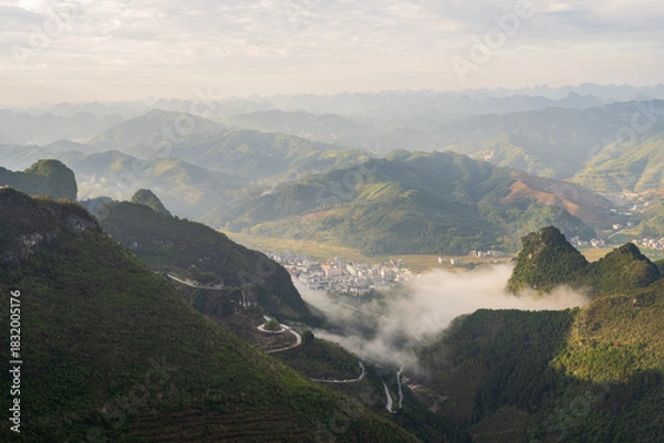Fototapeta Mountain Town Nestled in Valley With Drifting Clouds and Winding Roads in Guangxi China
