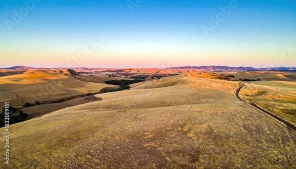 Fototapeta Aerial view of rolling hills covered in dry grass with a road winding through the landscape at sunset. The sky is a gradient of blue and orange.