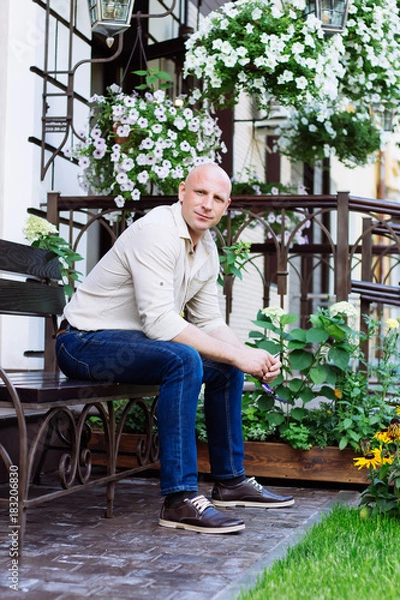 Fototapeta adult bald man sit on the bench at garden background at spring time. green grass, jeans, yellow flowers, jeans, shirt, boots. summer