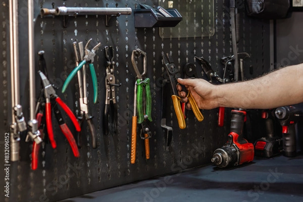 Fototapeta Mechanic's hand reaching for wire strippers on a pegboard wall. Organized workshop with wrenches, pliers, and impact driver.