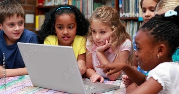 Obraz Leaning group of six students pointing at silver laptop on patterned table in library with shelves