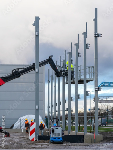 Fototapeta Workers installing steel support columns with telescopic handler at industrial construction site