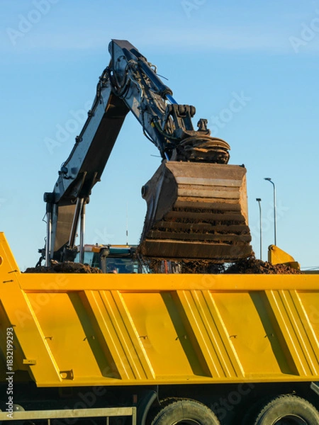 Fototapeta Excavator unloading soil into a yellow dump truck container at construction site on sunny day