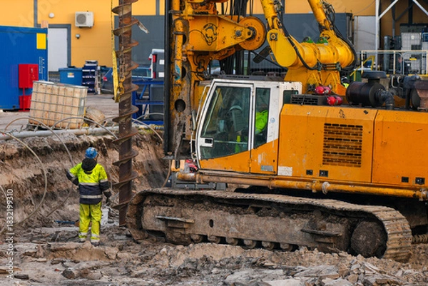 Fototapeta Heavy drilling machine operating at construction site with worker supervising foundation excavation