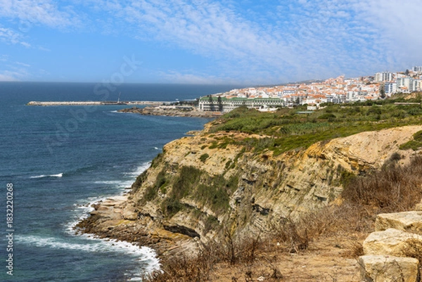 Fototapeta Stunning view of the Ericeira coastline in Portugal. The town's white buildings, including a large hotel, sit atop rugged cliffs by the blue sea.