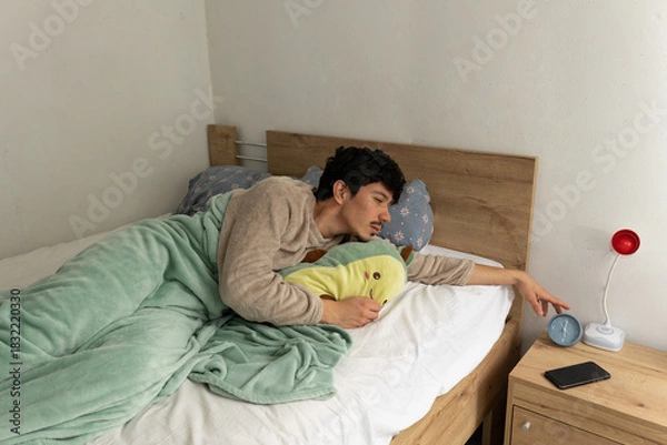 Obraz Man in pajamas, half asleep, turning off the alarm in the morning while slowly waking up in his bedroom. Concept of waking up and morning routine