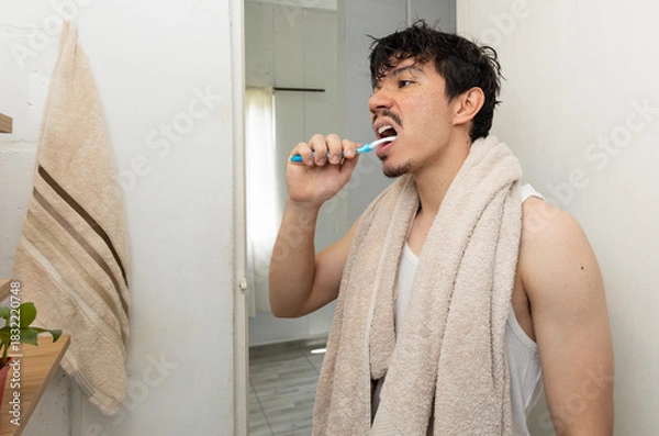 Obraz Man in the bathroom brushing his teeth with a towel around his neck, going through his morning hygiene routine. A scene of personal care and daily habits