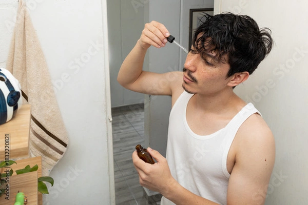 Obraz Man in the bathroom applying skincare with a dropper to his face as part of his morning facial care routine. A concept of wellness and self care at home