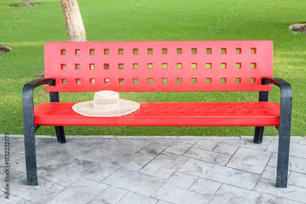 Fototapeta Red bench with straw hat in lush green park setting