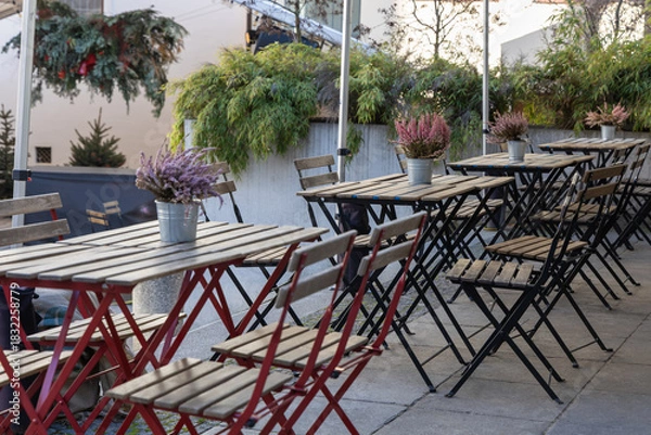 Fototapeta Outdoor seating area of a café with rows of folding wooden tables and chairs on a terrace. The tables are decorated with small tin buckets holding blooming flowering heath