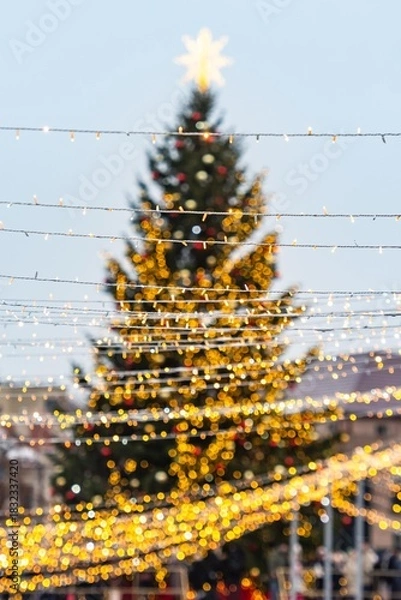 Fototapeta A large outdoor beautiful Christmas tree in Vilnius, decorated with warm white lights and ornaments, topped with a glowing star, standing in a festive setting with strings of lights in the foreground