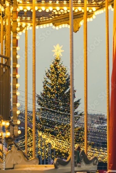 Fototapeta A large outdoor Christmas tree in Vilnius, decorated with warm white lights and ornaments, topped with a glowing star, standing in a festive setting with strings of lights in the foreground