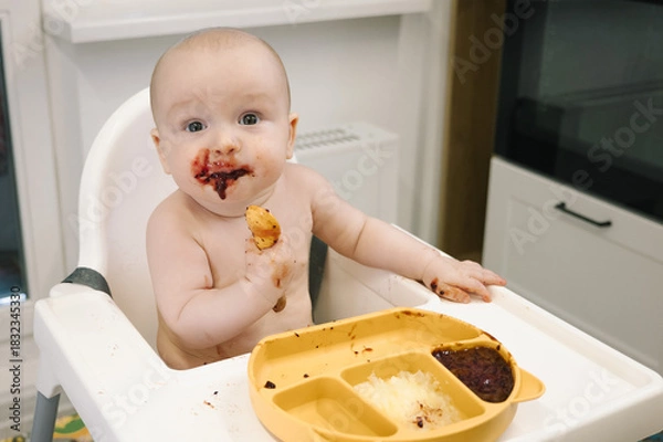 Fototapeta Toddler covered in food sits in a high chair and enthusiastically eating for meal on a spoon. Concept of early childhood, messy eating, and developmental milestones is conveyed.
