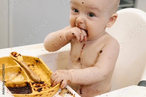 Fototapeta Toddler covered in food sits in a high chair and enthusiastically eating for meal. Concept of early childhood nutrition and introducing solid foods to infants.