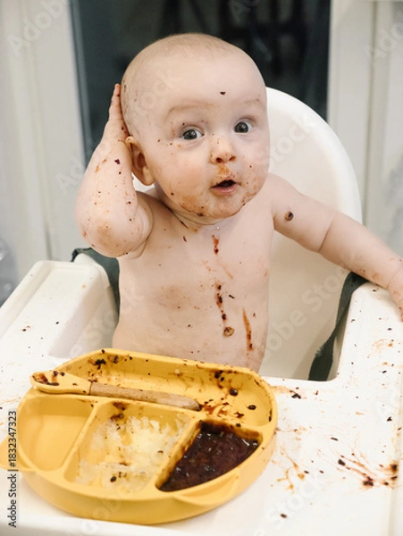 Fototapeta Toddler covered in food sits in a high chair and enthusiastically eating for meal. Concept of early childhood nutrition and introducing solid foods to infants.
