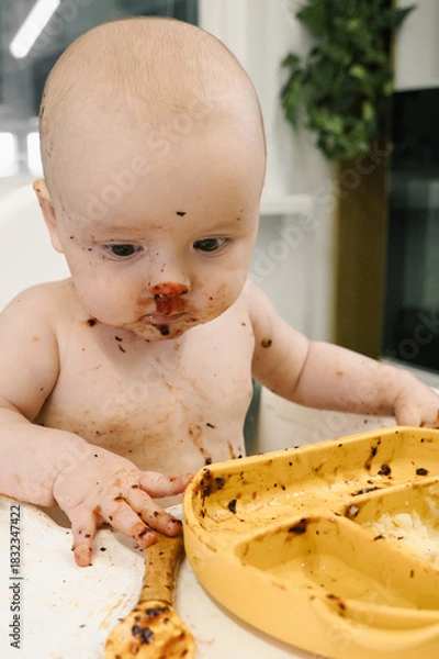 Fototapeta Toddler covered in food sits in a high chair and enthusiastically eating for meal. Baby playfully messy eating. Concept of early childhood nutrition and introducing solid foods to infants.