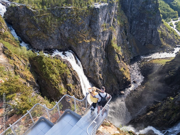 Fototapeta High-angle drone view of a couple on a modern viewing platform overlooking the majestic Voringsfossen waterfall in Norway.