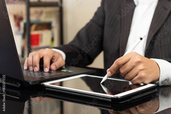 Fototapeta a CEO on suit at the right side of the screen writing something on a white tablet near to a laptop