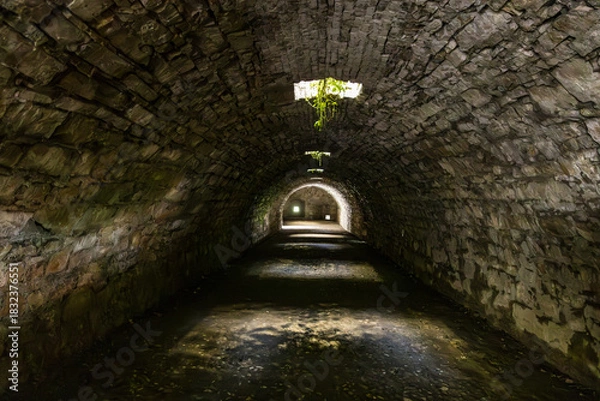 Fototapeta Interior view of one of the dark, vaulted stone tunnels or cisterns beneath the Castello di Brescia on Cidneo Hill, with light filtering through an overhead opening and illuminating the far end