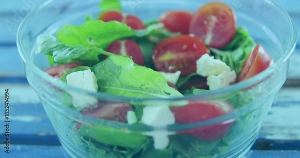 Fototapeta Showing clear glass salad bowl on painted blue table, with arugula, halved tomatoes, crumbled feta