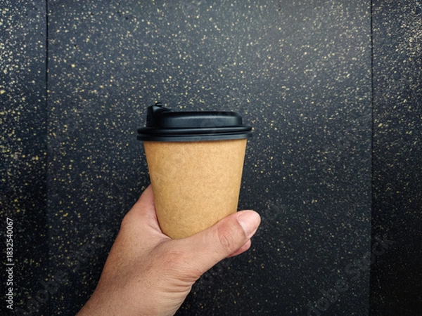 Obraz Hand holding a cup of coffee. Hand holding blank brown disposable paper coffee cup mockup with black lid and copy space for branding design.