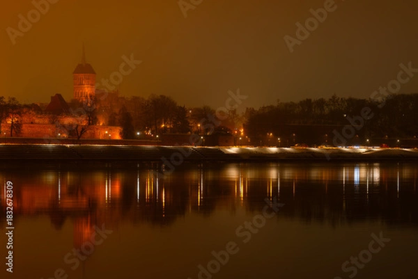 Obraz Toruń nocą, stare miasto nad rzeką Wisłą, światła odbijające się w wodzie. 