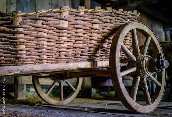 Fototapeta Vintage wooden cart with large wheels resting on a dusty floor in a rustic barn environment during the late afternoon light