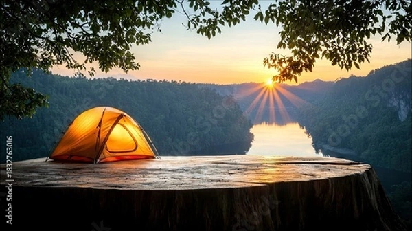 Fototapeta A camping tent illuminated from within sits on a large tree stump overlooking a river and mountains at sunset.